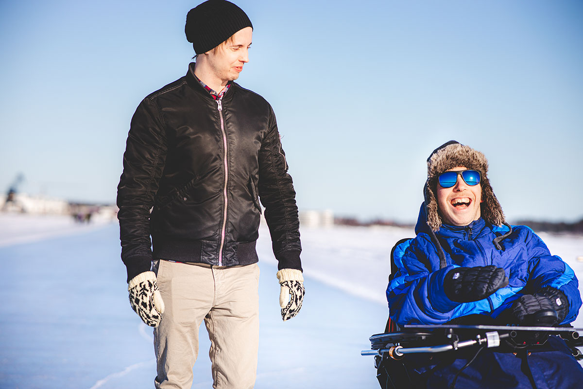 En leende kille i rullstol och en kille som går bredvid på isbanan i Luleå centrum