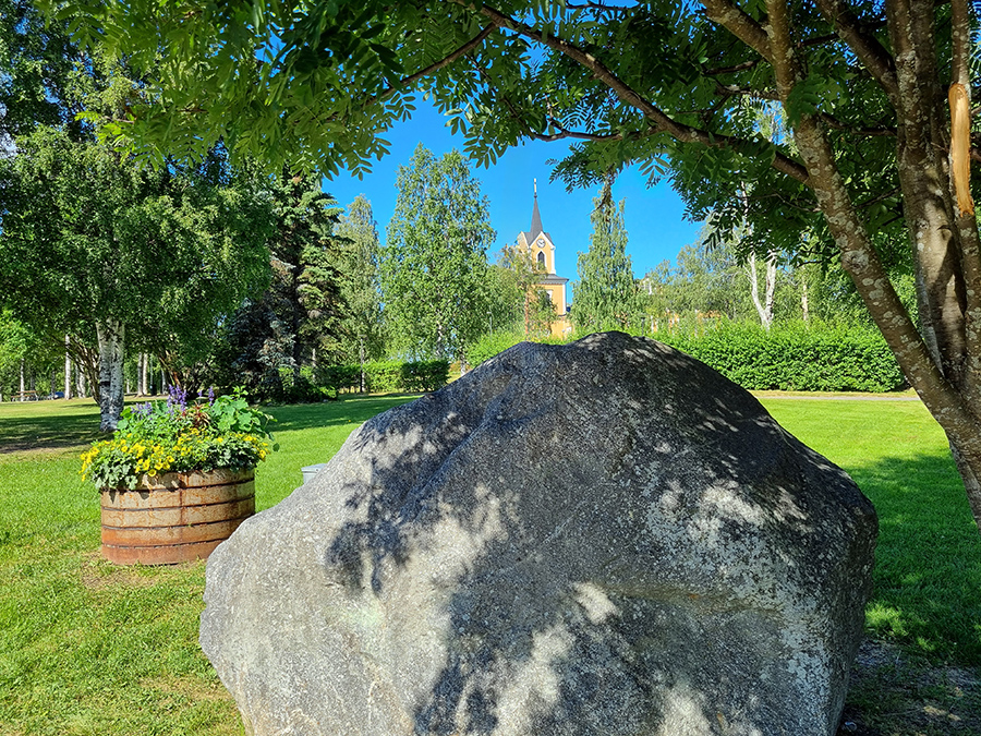Ett stort flyttblock ligger vid parkens utkant. I bakgrunden syns parken och Råneå kyrka.