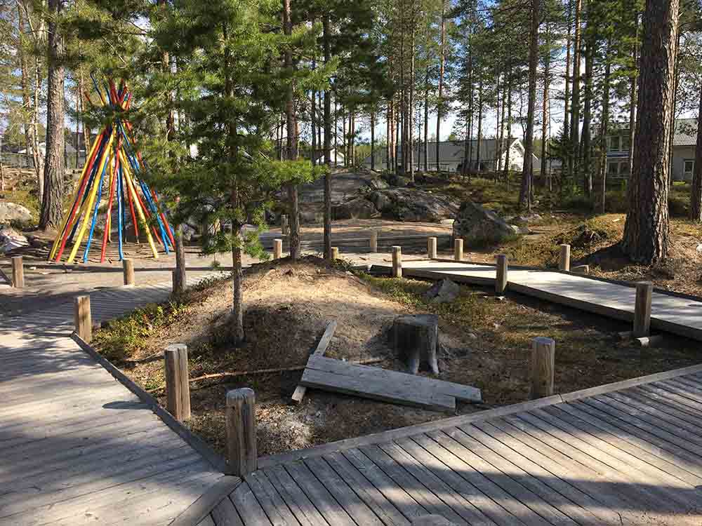 Träramper leder runt lekplatsen. Här har en yta lämnats mellan ramperna för lek. I bakgrunden synt färggrannar pinnar som står uppställda mot varandra så att de bildar en kåtaliknande konstruktion.