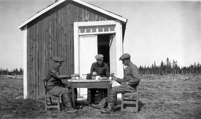 Tre män med kepsar och näbbstövlar sitter vid bord utanför en liten stuga.