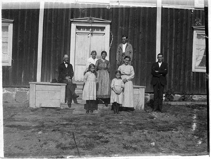 Familj framför ytterdörren till trähus. Tre män, tre kvinnor och två flickor.