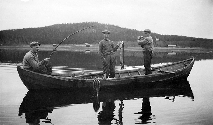 Tre unga män i eka på kav lugn sjö. En man håller upp en gädda, en annan håller i ett fiskespö.