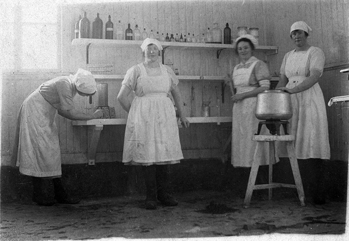 Fyra kvinnor med vita förkläden och mössor står framför bänk och vägghyllor med små glasflaskor på.