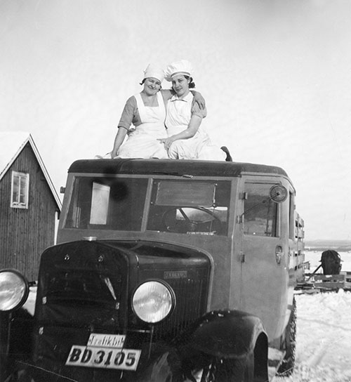 Två unga kvinnor med vita förkläden och mössor sittande på taket till en bil av äldre modell.