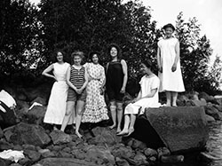 Sex kvinnor i badkläder och klänningar på stenig strand.