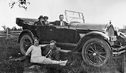 Bil av gammal modell med tre personer i bilen och en man och en kvinna i gräset vid sidan om.