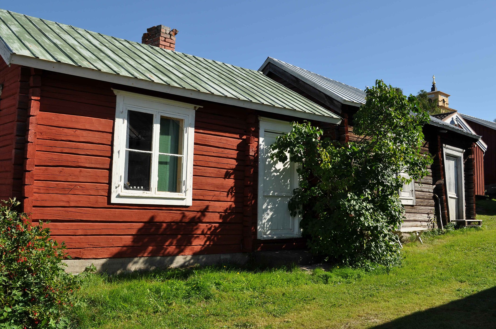 Röd liten timrad stuga med vitt fönster och vit dörr. Blå himmel, grön gräsmatta och träd jämte dörren.